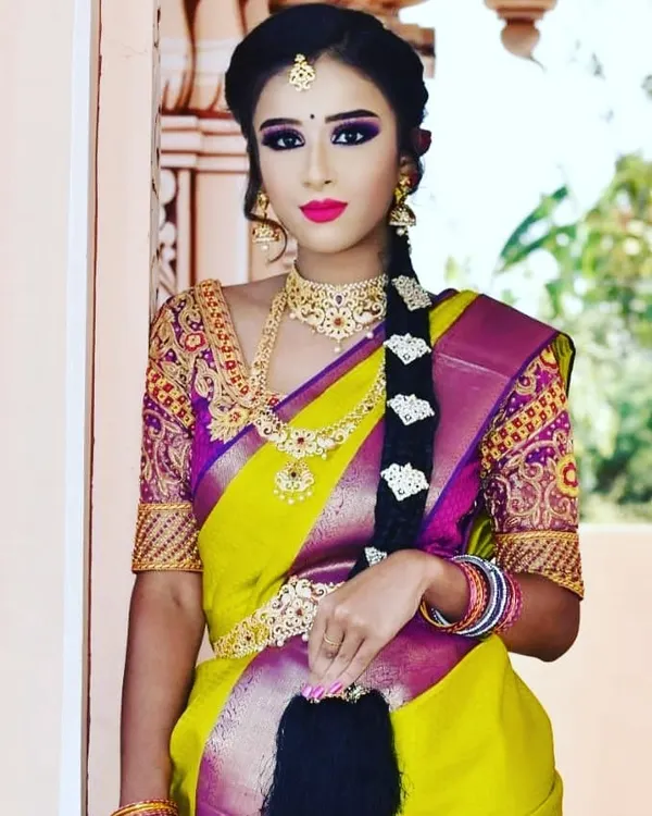 A full-length view of a traditional South Indian bridal style. The look features a classic long braid, a vibrant Kanjivaram saree, and makeup with a bold lip and defined eyes that tie the entire look together.