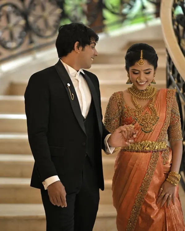 A classic and elegant look for the groom. This perfectly tailored black tuxedo with a satin lapel is a timeless choice that stands proudly next to the bride's traditional orange Kanjivaram saree.