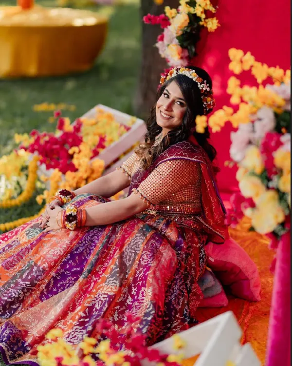 A beautiful, happy bride at her Haldi ceremony. She is wearing a vibrant, multi-colored lehenga and is adorned with a fresh flower crown, perfectly capturing the fresh and festive spirit of the occasion.