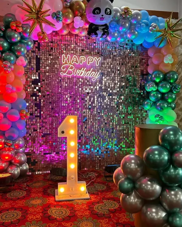 A vibrant first birthday party setup featuring a full balloon arch with a panda theme, a sparkling silver sequin wall, a "Happy Birthday" neon sign, and a large light-up marquee number '1'.