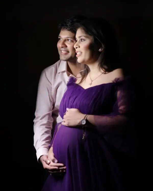 A classic low-key studio portrait of a couple. The lighting creates a warm, intimate mood, focusing on their happy expressions as they await their baby.