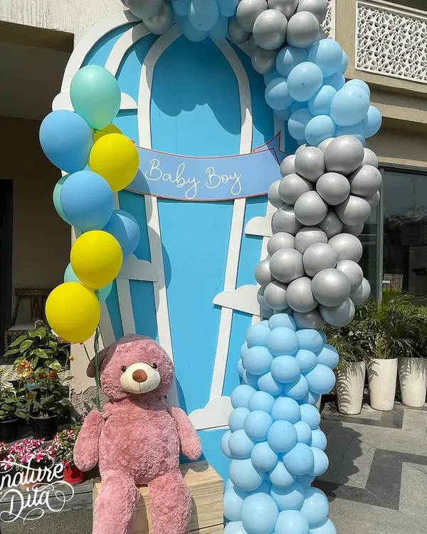 A beautiful 'Baby Boy' welcome setup for a baby shower, featuring a hot air balloon motif, a cascade of blue, yellow, and silver balloons, and a cuddly teddy bear.