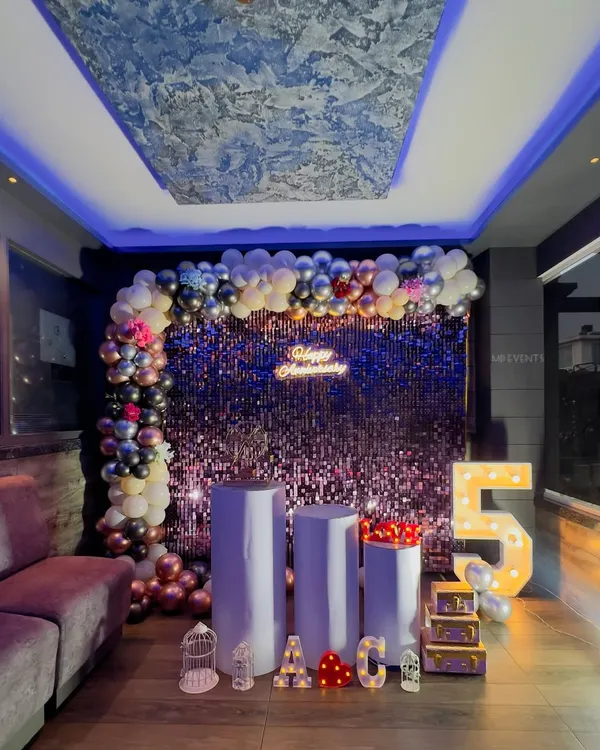 A glamorous 5th-anniversary party setup in a lounge. I used a purple shimmer wall, a multi-colored balloon garland, and large marquee numbers to create a vibrant and celebratory atmosphere.