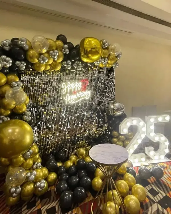A stunning 25th-anniversary decoration in black, gold, and silver. This setup includes a balloon arch, a silver sequin wall, a "Happy Anniversary" neon sign, and large marquee numbers.