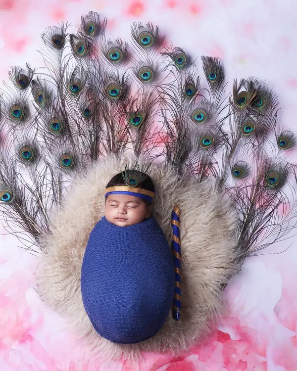 A creative Little Krishna theme for a newborn boy, featuring a comfortable blue wrap, a miniature flute, and a backdrop of real peacock feathers. This is one of my most requested artistic newborn setups.