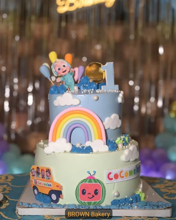 A colorful two-tier Cocomelon cake for a first birthday. This design features JJ, the iconic school bus, and a bright rainbow, creating a cheerful centerpiece for the little one's big day.