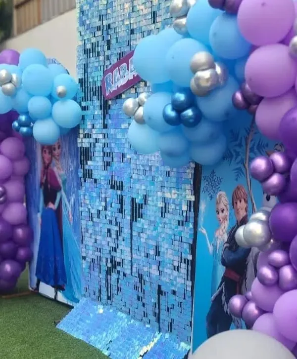 A close-up of our shimmering Frozen theme backdrop. The sequin wall and character cutouts of Elsa and Anna, framed by blue and purple balloons, create a magical icy palace.
