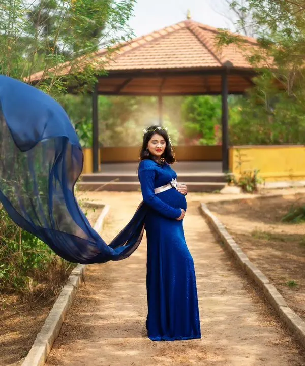 An expecting mother stands on a park pathway in a flowing royal blue maternity gown, the fabric caught in the wind to create a dramatic, wing-like effect. This outdoor maternity portrait captures a moment of power and beauty against a natural, sunlit backdrop.