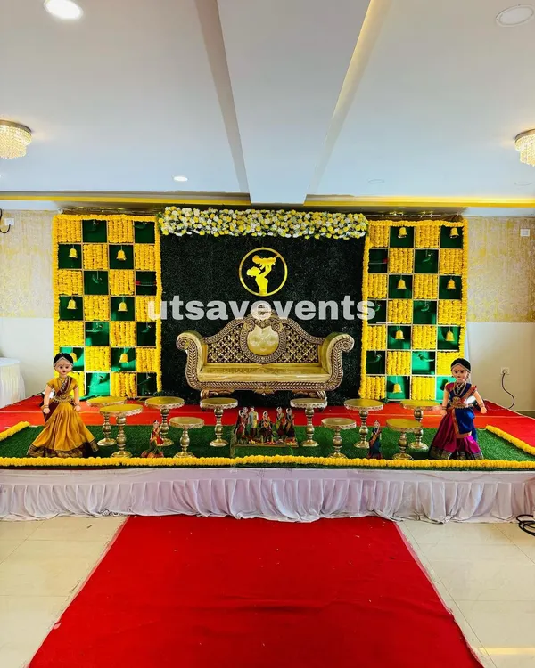 A classic Seemantham stage with a gold and green checkered marigold backdrop. The setup is completed with a traditional divan, brass urlis, and Marapachi dolls for an authentic feel.