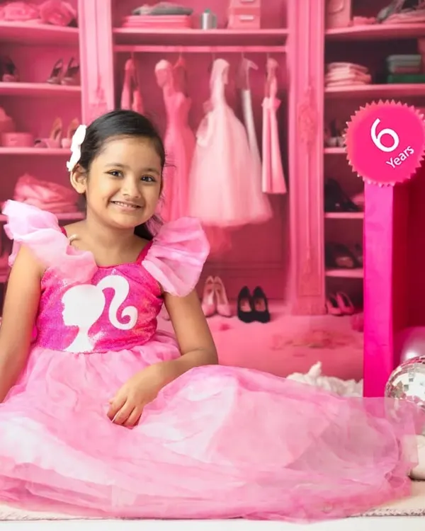 A six-year-old girl smiles brightly in her Barbie-themed shoot, sitting beside a life-sized Barbie box in a room filled with pink.