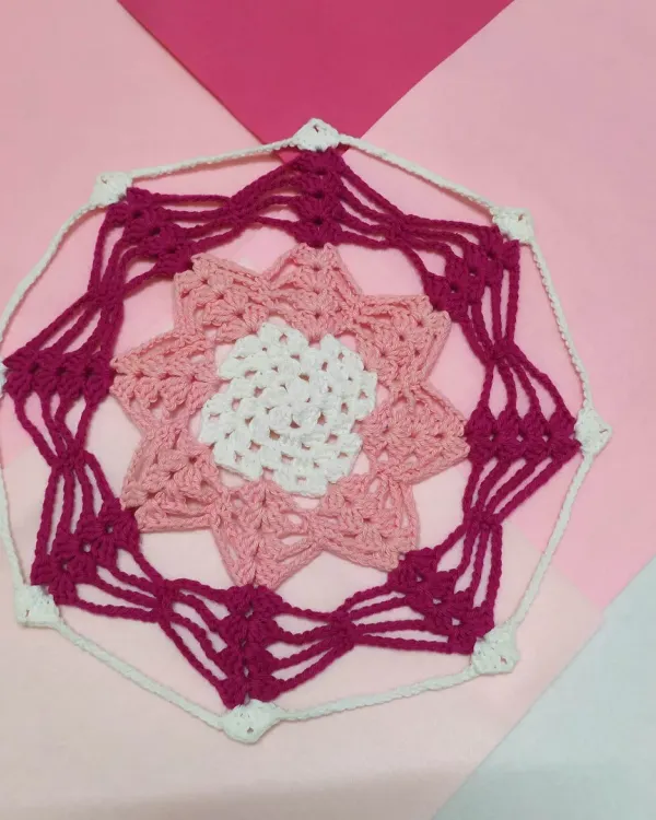 This is the Karamella doily, an intricate design featuring layers of stitches in shades of pink and white. It can be used to decorate a table or hung as a beautiful piece of wall art.