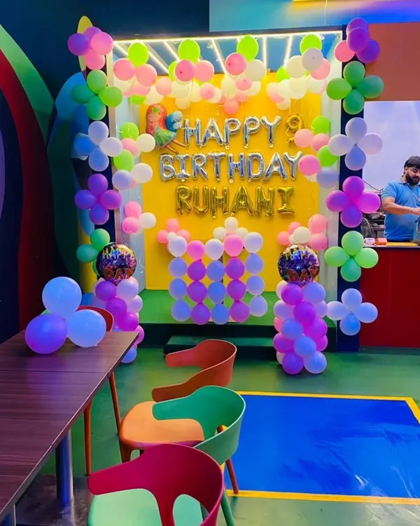 A colorful birthday setup in a cafe for Ruhani's 9th birthday. The decor features a bright yellow backdrop with a pastel balloon garland and "Happy Birthday" foil letters.