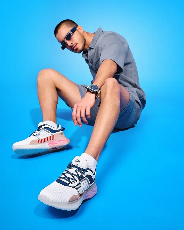 A modern shoe campaign featuring a model in a casual pose against a vibrant blue background. The wide-angle perspective emphasizes the stylish sneakers, making them the focal point of this commercial shot.