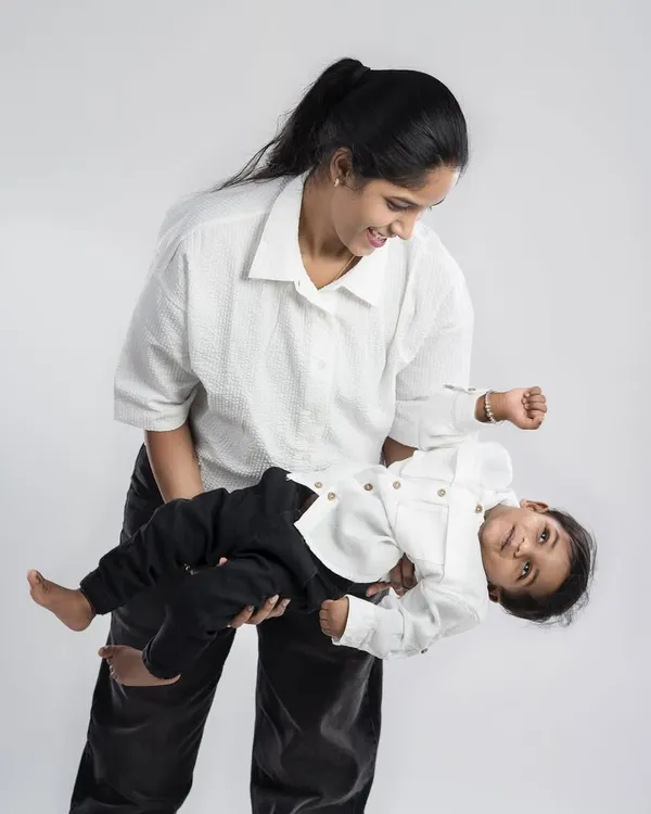 A mother holds her baby boy, their matching white shirts creating a clean and simple look. The playful tilt and their smiles capture a moment of pure, uncomplicated joy.