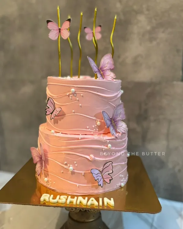 A beautiful two-tier butterfly themed cake. The soft pink buttercream has a gentle wave texture, adorned with delicate butterflies and pearl sprinkles, making it perfect for a special birthday.