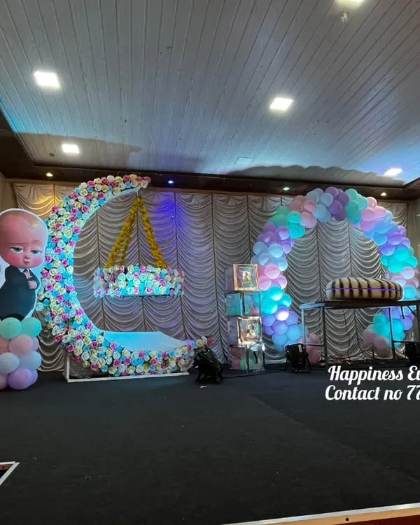 This naming ceremony stage was designed with a Cocomelon theme. It features a large floral crescent moon with a hanging cradle, a balloon arch, and cutouts of the popular Cocomelon characters.