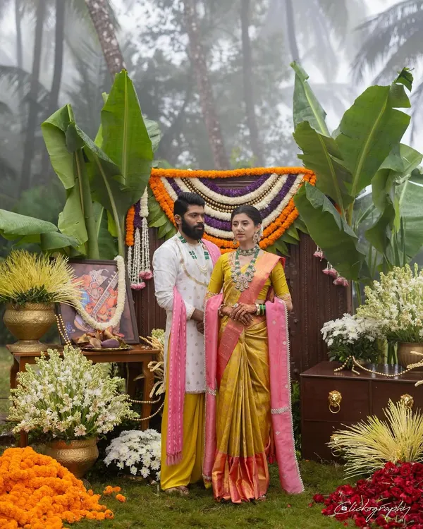 A beautifully coordinated couple on their wedding day, set against a stunning backdrop of traditional floral decor. This look captures the essence of a vibrant, modern Maharashtrian wedding, blending a classic yellow Paithani saree with a contemporary pink shela and a custom-designed groom's outfit.
