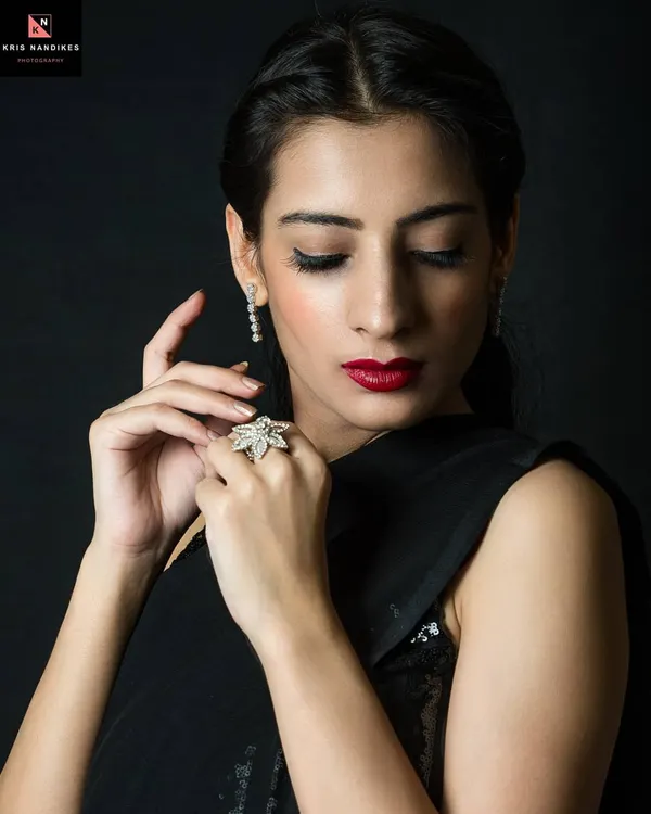 An elegant low-key portrait of a woman in a black saree. The lighting gently illuminates her face and hands, highlighting her red lipstick and diamond jewelry against the dark background.