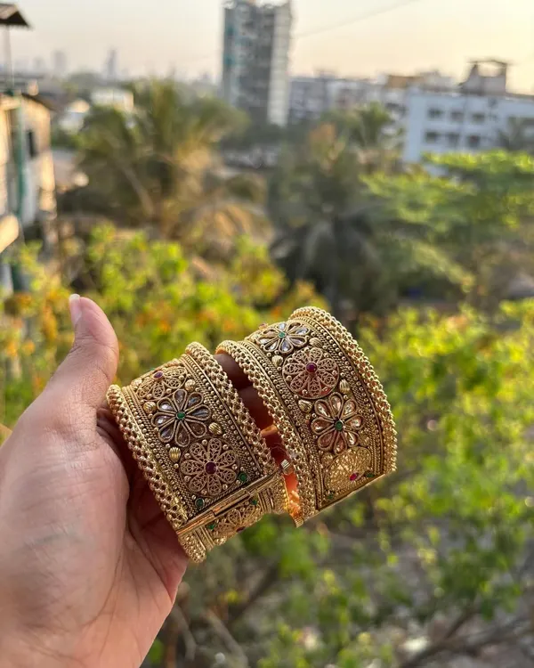 A pair of our premium collection bangles, held up against the light. These broad kadas feature intricate floral designs with red and green stones.