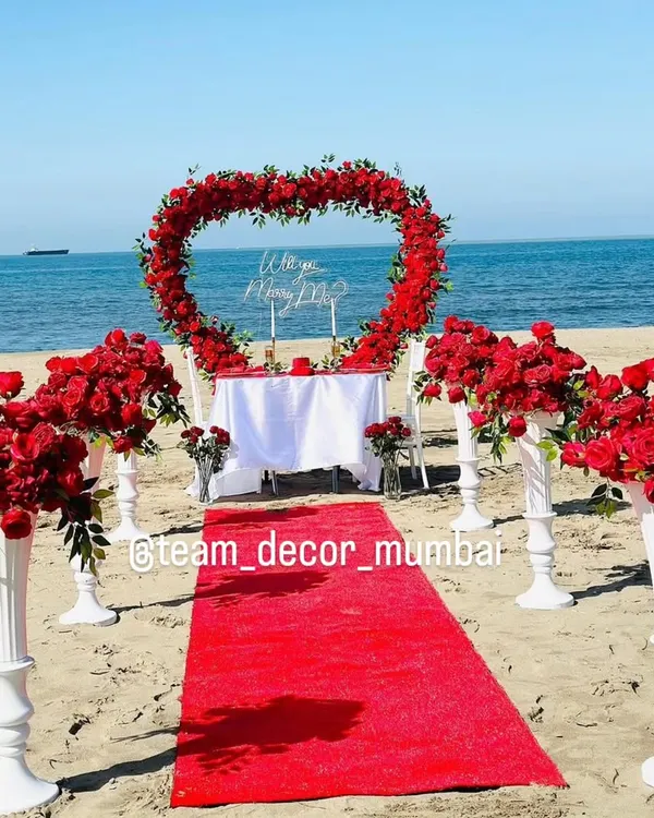 A full view of the romantic beach proposal setup I offer in Mumbai. It includes a red carpet aisle, a heart-shaped arch of red roses, floral pedestals, and a beautifully set table for two, right by the sea.