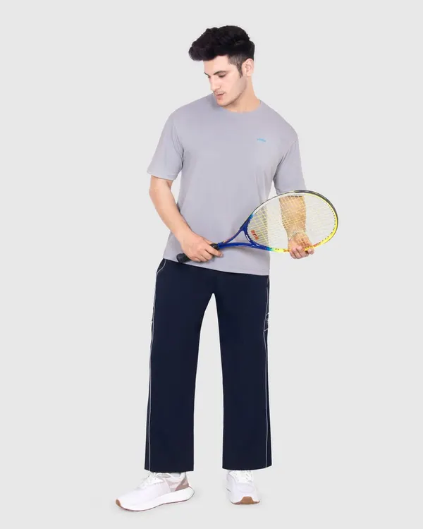 A full length e-commerce model shot for a sportswear brand. The clean grey background ensures no distractions, keeping the focus entirely on the fit and style of the t-shirt and track pants.