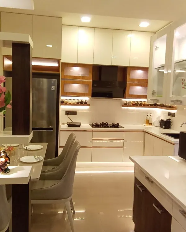 This L-shaped modular kitchen is designed for both beauty and function. The combination of off-white and wood-finish cabinets, frosted glass shutters, and under-cabinet lighting creates a warm, inviting space for cooking and gathering, complete with a cozy breakfast counter.
