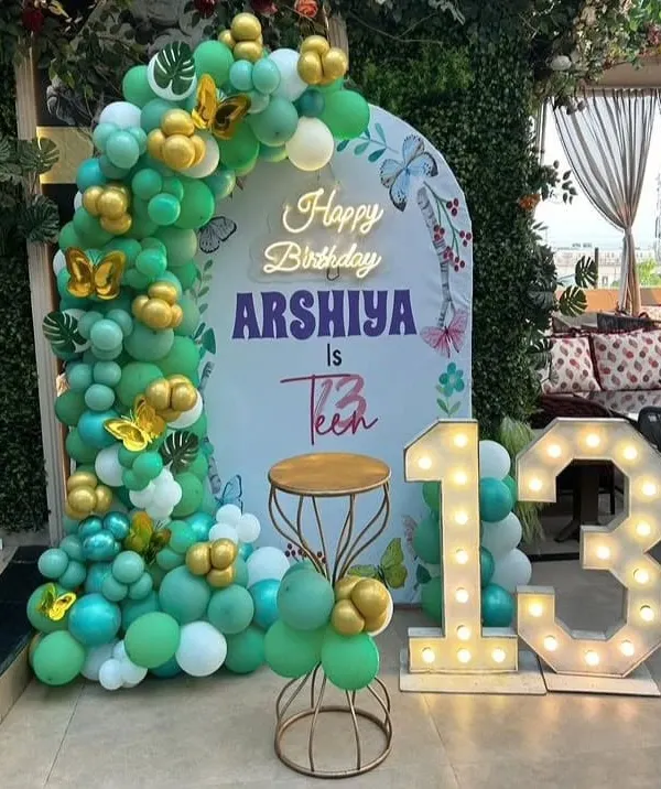 A beautiful butterfly theme for a 13th birthday. This outdoor setup features a teal and gold balloon arch, a personalized backdrop, and large light-up numbers for the big teen birthday.