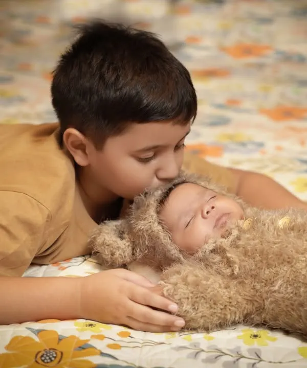 A moment of pure tenderness between brothers. An older brother lies on a floral patterned floor and gives a gentle kiss to the forehead of his baby sibling, who is sleeping peacefully in a fuzzy bear costume.