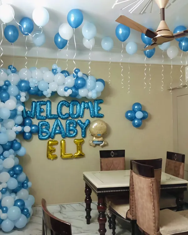 A classic "Welcome Home" decoration for a baby boy. This living room setup features a large organic balloon arch in shades of blue and white, with foil letters spelling out "Welcome Baby Eli" to make the moment unforgettable for the new parents.