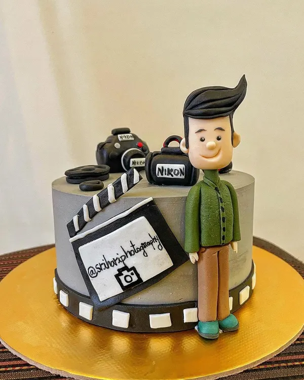 The perfect cake for a photographer! This design features a hand-sculpted fondant caricature, a miniature Nikon camera, a clapperboard, and a film strip border, celebrating a passion for capturing memories.