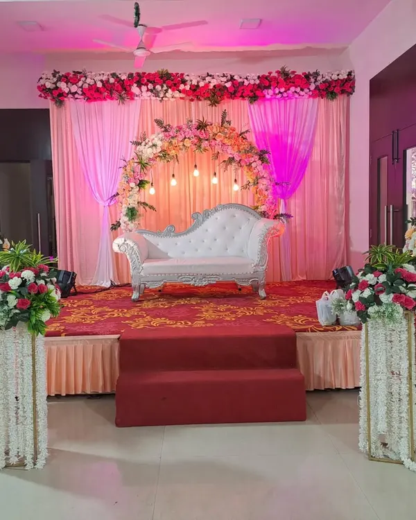An elegant stage setup for a reception or engagement, featuring a white chaise lounge against a backdrop of pink curtains. The look is completed with a floral arch and matching flower-wrapped pillars.