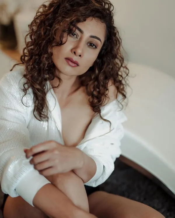 A soft and intimate indoor portrait. The model's expressive eyes and naturally curly hair are the focus, framed by a simple white sequined shirt and gentle lighting.