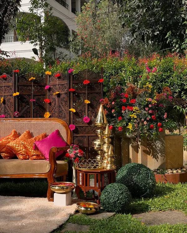 A beautiful outdoor seating area for a Mehandi function. The setup features a traditional carved wooden screen, vibrant pink and orange cushions, and brass decor, creating a rich and colorful ambiance.