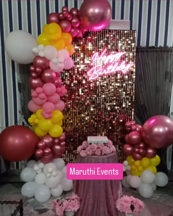 A glamorous birthday setup featuring a gold shimmer wall, a 'Happy Birthday' neon sign, and a colorful balloon arch in shades of pink, yellow, and white.