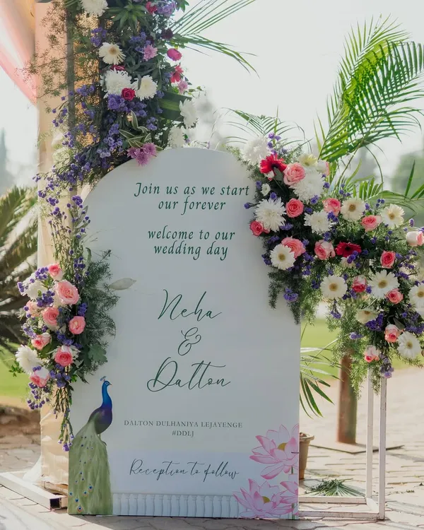 A beautiful welcome sign for a wedding, designed in a modern arch shape with elegant calligraphy. We adorned it with lush arrangements of pastel flowers and a peacock illustration to add a touch of Indian royalty.