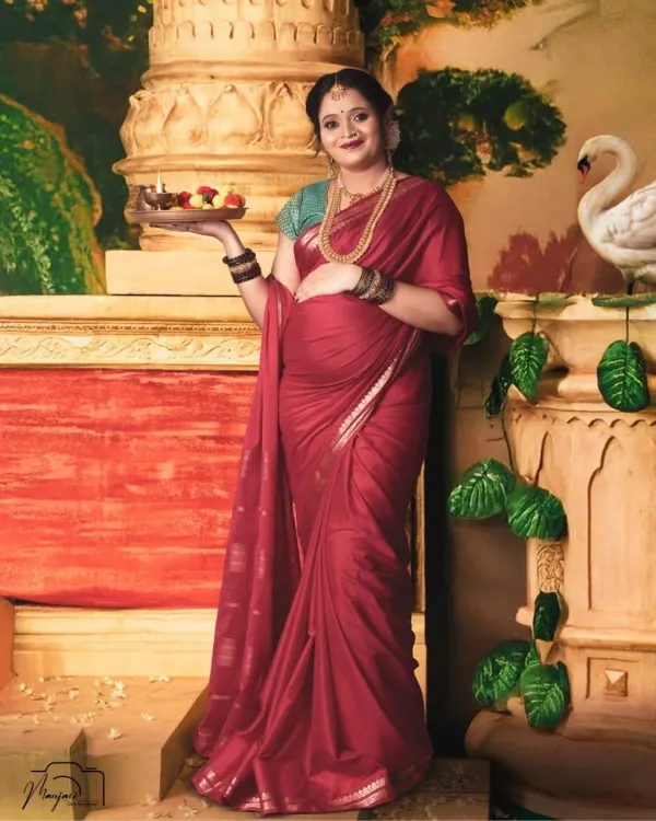 A vision of traditional grace. This mom-to-be, dressed in a beautiful red saree and holding a puja thali, is captured in a setting that evokes the feeling of a classic Indian painting.