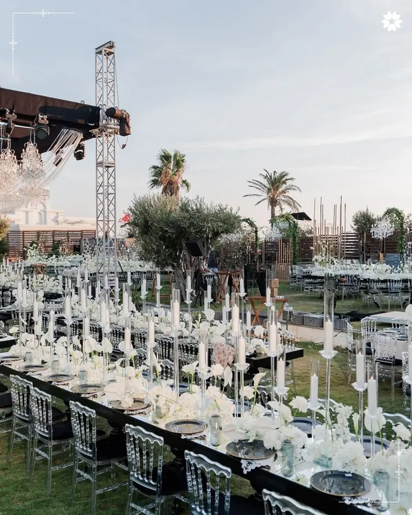 An outdoor reception with long tables adorned with a sea of candles and white florals. The modern, transparent chairs and dramatic chandeliers create a stunning contrast against the open-air setting, crafting an unforgettable dining experience.