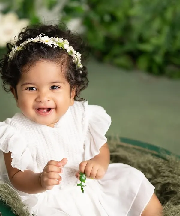 A beautiful sitter session portrait with a lush green backdrop. Her bright, happy smile and the simple white dress make this a timeless and classic baby photo.