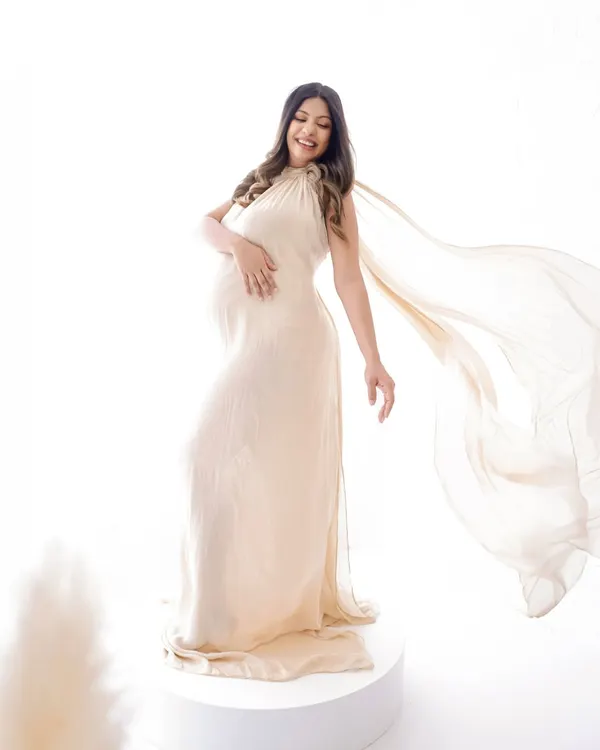 A moment of pure joy and elegance. This mama-to-be is captured in a flowing cream gown against a high-key white background, creating a light and ethereal portrait.