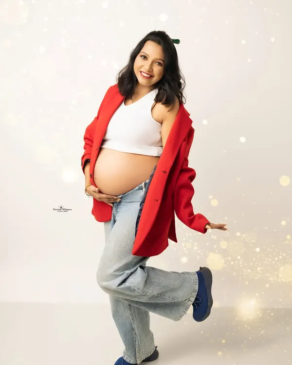 A fun and festive Christmas-themed solo portrait. The mother-to-be is dressed in a cheerful red blazer and jeans, kicking up her heel with joy.