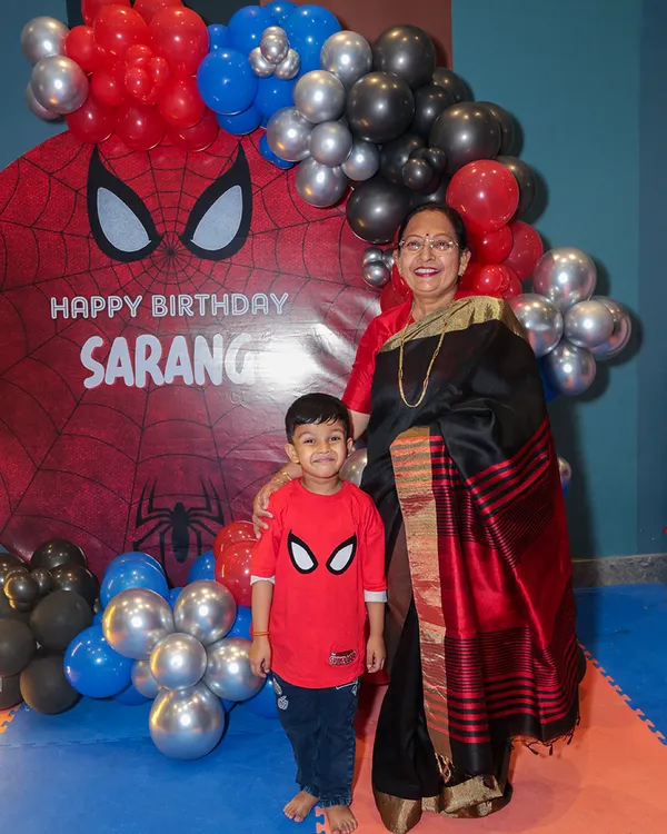 The birthday boy, dressed in his Spiderman shirt, stands proudly with his grandmother in front of the custom superhero backdrop, a perfect photo opportunity.