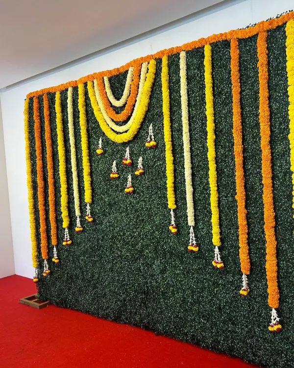 A versatile greenery wall backdrop. This simple yet elegant design is decorated with hanging garlands of yellow, orange, and white flowers, suitable for any festive occasion.