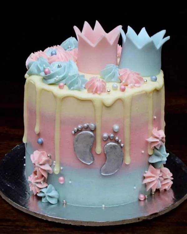 A sweet baby shower cake perfect for a gender reveal party. It features a pink and blue ombre design, a white chocolate drip, and is topped with little pink and blue fondant crowns. The tiny silver baby footprints are my favorite part.