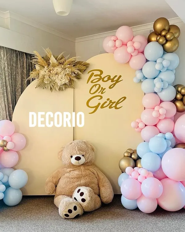 A classic "Boy or Girl" gender reveal party setup. This design uses a neutral backdrop with gold lettering, a large teddy bear, and a mix of pink and blue balloons to build anticipation for the big reveal.