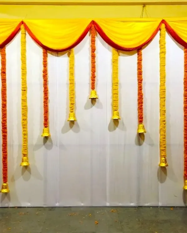 A simple and traditional Haldi ceremony backdrop for home. The decor uses yellow and orange marigold garlands with temple bells hanging from a yellow fabric drape.