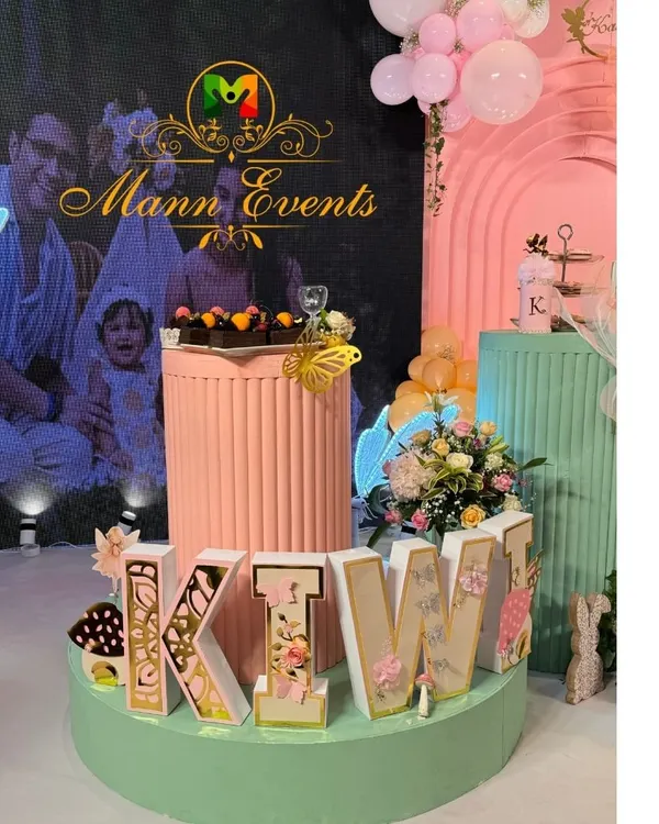 A close-up of a pastel-themed dessert table, featuring personalized 'KIWI' letters, a neon butterfly, and a photo of the family on the LED screen. The details make the difference.