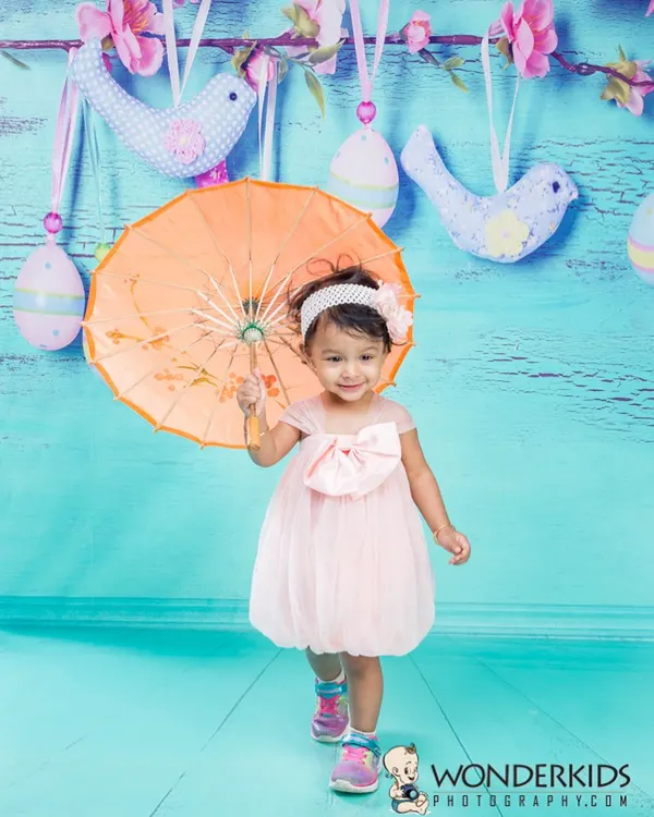 A sweet toddler in a pink dress holding a bright orange umbrella. The colorful props and cheerful blue background create a very happy and playful portrait.