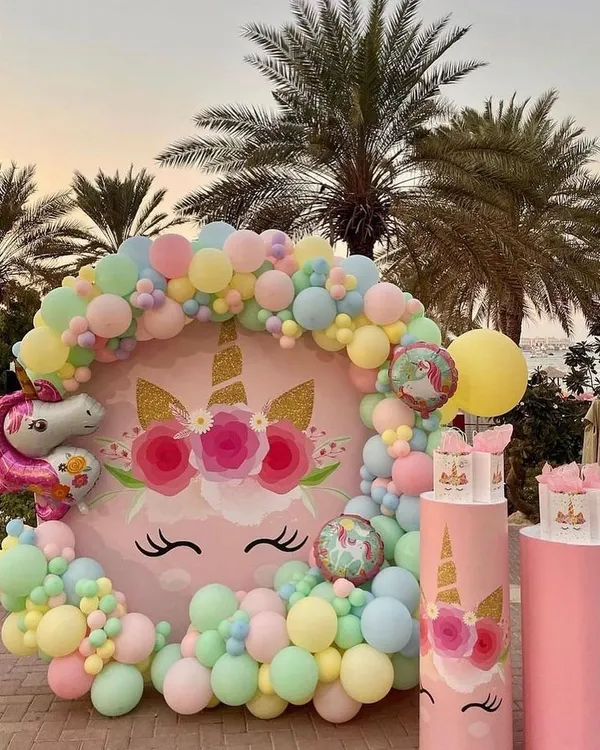A beautiful outdoor unicorn party setup with a circular backdrop featuring a unicorn face, surrounded by a delicate pastel balloon garland. Themed plinths are ready for the cake and treats.