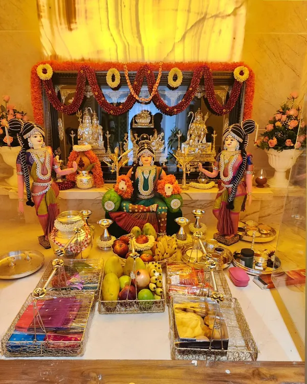 A complete Varamahalakshmi pooja setup with the goddess seated on a throne, surrounded by fruits, flowers, and traditional offerings. The decor includes a beautiful flower *thorana* in the background.