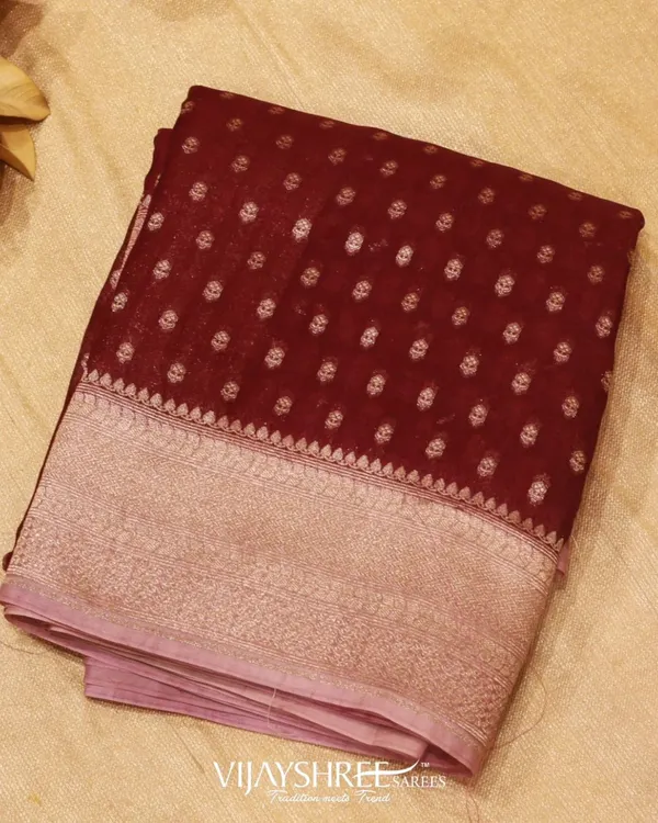 A dark maroon Kora silk saree adorned with beautiful floral motifs and a grand silver zari border. Kora silk is known for its texture and lightweight feel, offering a graceful drape.